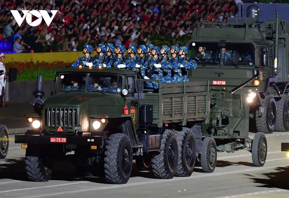 The soldiers, in solemn composure, ride atop the towed anti-aircraft guns as they roll past the square.