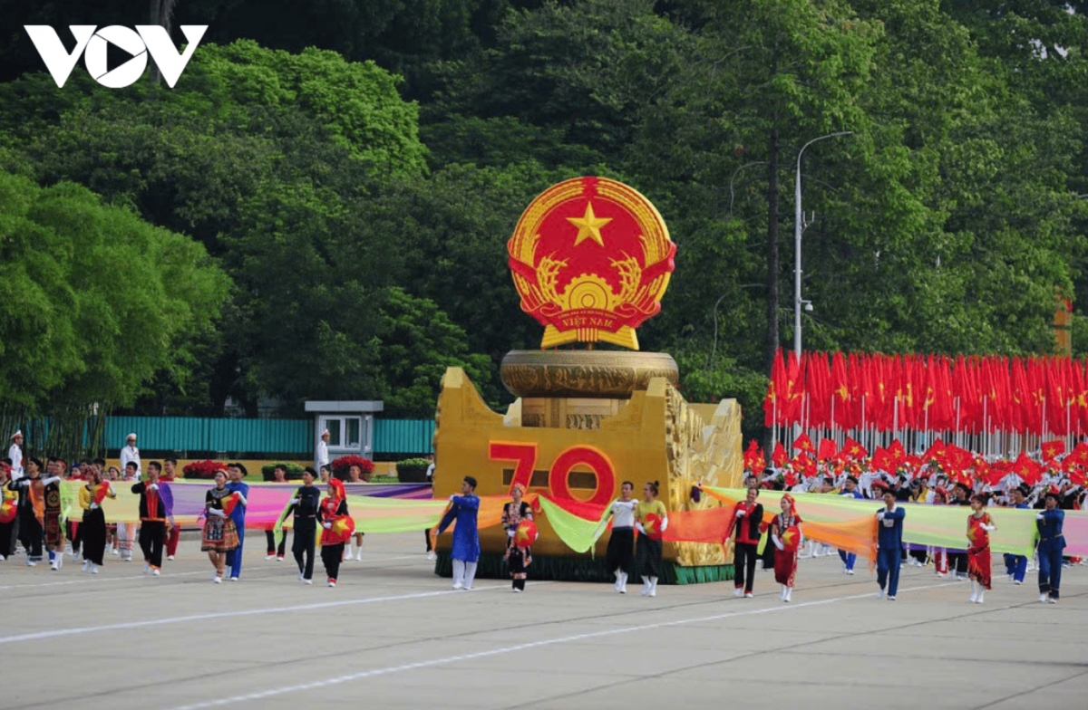 The parade is led by the ceremonial vehicle displaying the National Emblem, followed by a formation carrying the Party flag and the National flag.